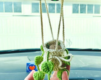 May include: A handmade, cream-colored crochet plant holder with a wooden bead accent, suspended by cream-colored rope. The holder contains a small, green, crochet plant with heart-shaped leaves. The plant is hanging in a car.