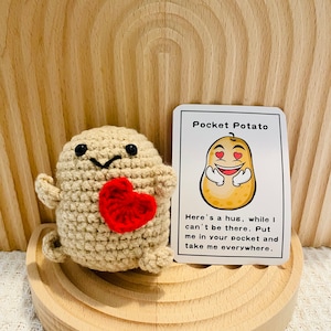 May include: A crocheted potato toy holding a red heart. The toy is sitting on a wooden surface with a card that reads "Pocket Potato" and "Here's a hug, while I can't be there. Put me in your pocket and take me everywhere."