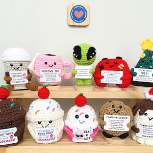 May include: A collection of handmade crochet items, including coffee cups, cupcakes, and a Christmas tree. Each item has a smiling face and holds a sign with positive messages. The color palette includes brown, pink, green, and white.