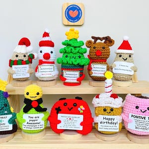 May include: A collection of handmade crochet figures, including a snowman, Christmas tree, reindeer, and cupcake, each holding a sign with positive messages. The figures are displayed on a wooden shelf, with a heart-shaped logo in the background.