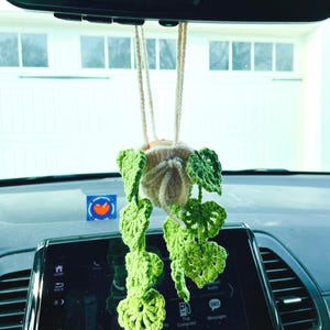 May include: A handmade crochet plant car charm with green leaves and a beige pot. The charm hangs from the rearview mirror, adding a touch of nature to the car's interior. The leaves are a vibrant green color.
