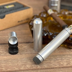 Puede incluir: Dos tubos de cigarros plateados con detalles negros. Un tubo tiene una tapa negra con una malla de metal. El otro tubo tiene una punta de espuma negra. Los tubos están sobre una superficie de madera.