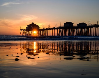 May include: Silhouette of a pier with a building at the end, the setting sun shining through the structure, reflecting in the water below.
