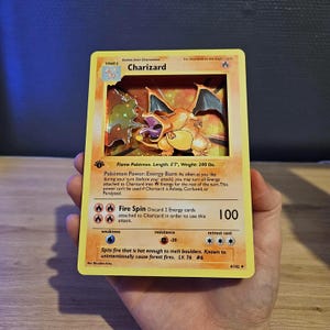 Peut inclure: Une carte à collectionner en relief 3D représentant le personnage Charizard. La carte présente une bordure jaune, un fond orange, le nom Charizard, le niveau Stage 2, Flame Pokémon, taille 170 cm, poids 91 kg et l'attaque Fire Spin à 100 dégâts.
