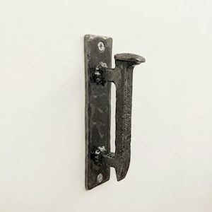 May include: A rustic, dark metal hook mounted on a rectangular plate. The hook is shaped like a large nail, with a textured surface and visible hammer marks. Two screws secure the plate to a white surface, suggesting a wall-mounted design.