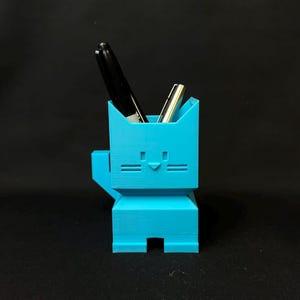 May include: A bright turquoise cat-shaped pen holder with two pens inside. The holder has a geometric design with a square body, a small arm, and a face with horizontal lines for the eyes and mouth. The pens are black and silver.