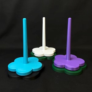 May include: Three thread spool holders, each with a flower-shaped base and a central post. The holders are in blue, white, and purple, with layered bases in black and green. These are designed to hold and organize thread spools, ideal for sewing.