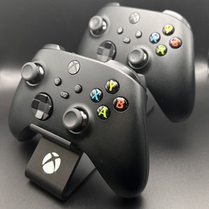 May include: Two black Xbox controllers with coloured buttons, one on a charging stand. The stand has a white Xbox logo.