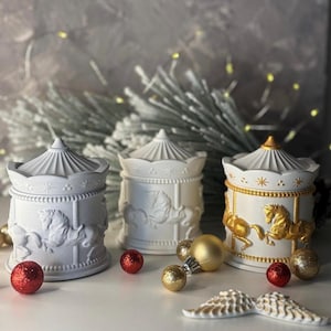 Op de afbeelding: Drie carrouselvormige decoratieve dozen in wit en goud. De dozen hebben carrouselpaardenontwerpen en zijn versierd met gouden en rode accenten. De scène is geplaatst met feestelijke ornamenten en een besneeuwde achtergrond.