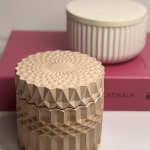 Puede incluir: Dos portavelas decorativos de hormigón. Uno es blanco con crestas verticales y el otro es marrón claro con un patrón geométrico.