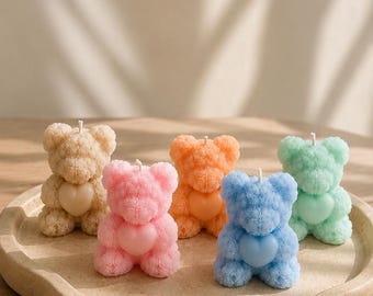 May include: Five bear-shaped candles with textured, fuzzy-looking surfaces, each holding a smooth heart on its chest. The candles are colored beige, pink, orange, blue, and mint green, and each features a single white wick at the top of its head.