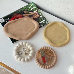 May include: Four decorative trays in different shapes and colors. The trays are made of a smooth, textured material. One tray is a light brown, irregular shape. Another tray is a light yellow, irregular shape. The third tray is a white, round tray with a beaded edge. The fourth tray is a light pink, round tray with a beaded edge. The trays are perfect for organizing small items like jewelry, keys, or coins.