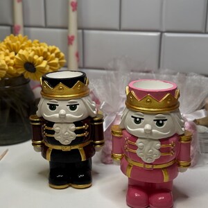 Puede incluir: Dos figuras decorativas de cascanueces. Una es negra y burdeos con una corona dorada, la otra es rosa con una corona dorada. Ambas tienen cabello blanco y ojos verdes. El fondo incluye velas y flores.