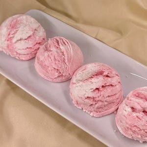 May include: Four pink and white bath bombs arranged in a row on a white rectangular plate. The bath bombs are shaped like ice cream scoops.