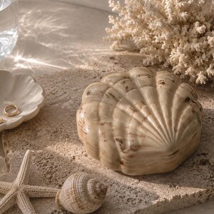 May include: A beige and brown marbled shell-shaped trinket box with a ridged surface. The box is positioned alongside a small white shell dish containing a gold ring, a beige starfish, a small spiral seashell, and a piece of white coral.