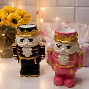 May include: Two decorative nutcracker figurines. One is black with gold accents, the other is pink with gold accents. Both have white hair, green eyes, and gold crowns. They are standing upright on a white surface.