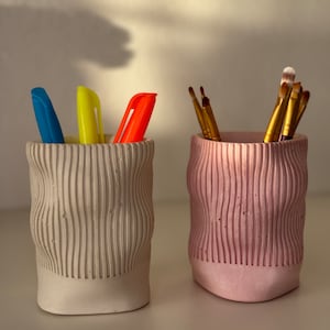 May include: Two textured ceramic pencil holders, one in a light gray color and the other in a light pink color. The gray holder has three pens inside, one blue, one yellow, and one orange. The pink holder has several paintbrushes inside.