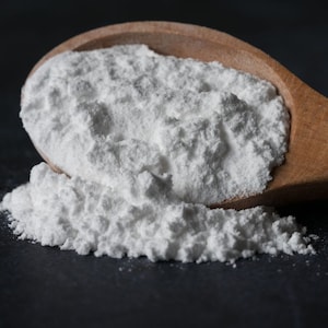 Könnte beinhalten: Ein Holzlöffel gefüllt mit weißem Backpulver, das auf eine schwarze Oberfläche rieselt.