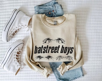 May include: A beige sweatshirt with the text "batstreet boys" in black, accompanied by bat wing graphics. The sweatshirt is paired with light-wash denim jeans and white high-top sneakers. A gold chain necklace is visible.