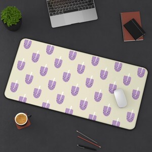 May include: A rectangular desk mat with a repeating pattern of purple bubble tea cups on a cream background. The mat is surrounded by a black border. A white computer mouse is on the mat.