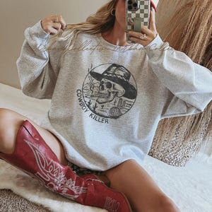 Può includere: Una felpa grigio chiaro con una grafica circolare raffigurante un teschio con un cappello da cowboy, che fuma una sigaretta, e le parole "Cowboy Killer". La persona indossa un cappellino da camionista rosso e bianco con la scritta "Timeless" e stivali da cowboy rossi.