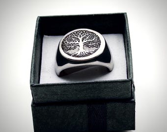 May include: A silver-colored ring with a detailed tree design set in a circular frame. The ring is displayed inside a black gift box with a white interior. The ring's band is thick and polished.