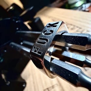May include: A silver ring with the word "FUCK" engraved on the band. The ring is held by a tool, with a wooden surface in the background. The ring is made of a shiny metal.