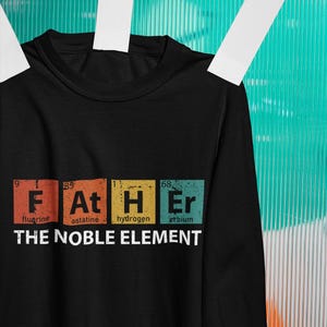 Puede incluir: Camiseta negra de manga larga con la palabra "FATHER" escrita con elementos de la tabla periódica. El texto "THE NOBLE ELEMENT" está debajo. Las letras son de color naranja, amarillo y verde azulado.