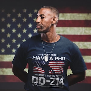 Può includere: T-shirt blu navy con la scritta "I MAY NOT HAVE A PHD BUT I HAVE A DD-214" su un disegno della bandiera americana. Lo sfondo presenta la bandiera americana. La persona indossa una collana con un'etichetta d'argento.