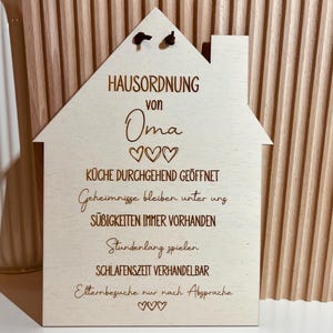 May include: A wooden house-shaped sign with the text "HAUSORDNUNG von Oma" and hearts. The sign lists house rules, including "K&uuml;che durchgehend ge&ouml;ffnet", "Geheimnisse bleiben unter uns", "S&uuml;&szlig;igkeiten immer vorhanden", "Stundenlang spielen", "Schlafenszeit verhandelbar", and "Elternbesuche nur nach Absprache".