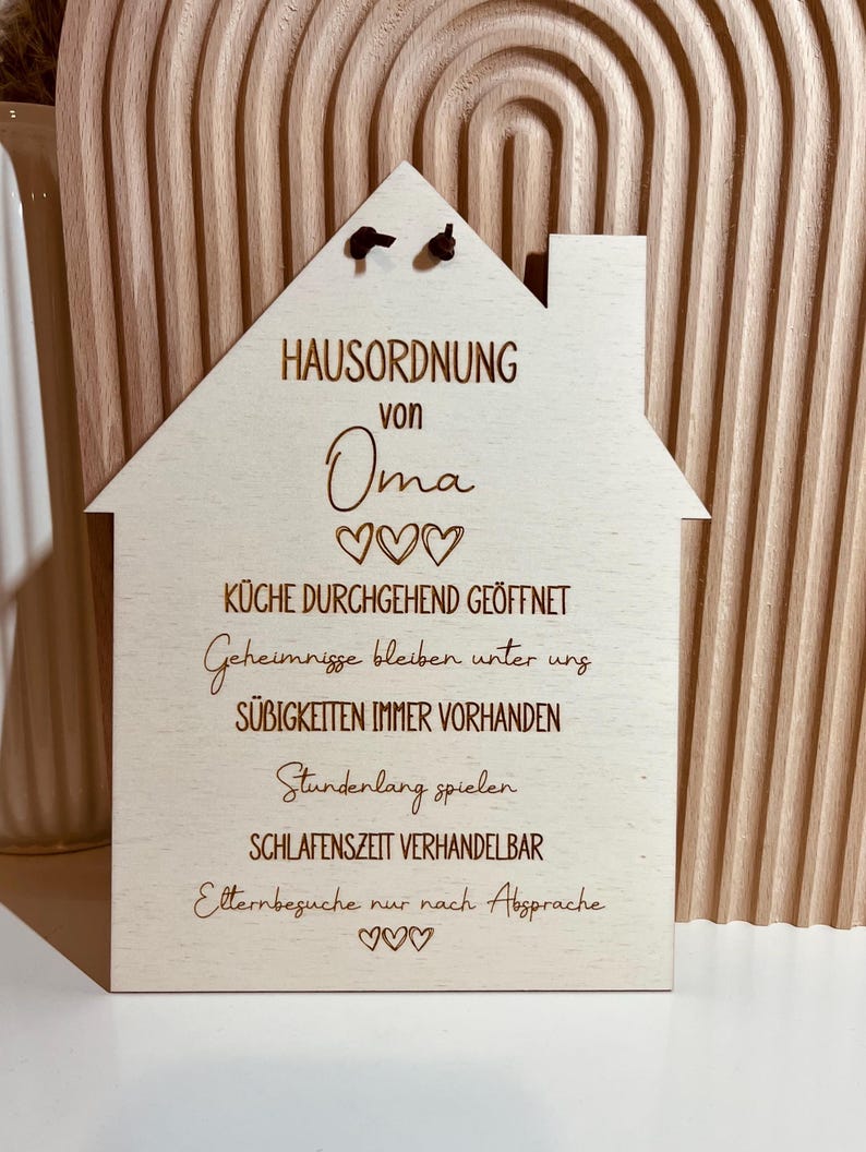 May include: A wooden house-shaped sign with the text "HAUSORDNUNG von Oma" and hearts. The sign lists house rules, including "K&uuml;che durchgehend ge&ouml;ffnet", "Geheimnisse bleiben unter uns", "S&uuml;&szlig;igkeiten immer vorhanden", "Stundenlang spielen", "Schlafenszeit verhandelbar", and "Elternbesuche nur nach Absprache".
