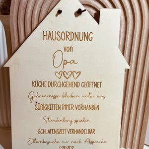 May include: A wooden house-shaped sign with the text "HAUSORDNUNG von Opa" and rules for the house, including "K&uuml;che durchgehend ge&ouml;ffnet", "Geheimnisse bleiben unter uns", "S&uuml;&szlig;igkeiten immer vorhanden", "Stundenlang spielen", "Schlafenszeit verhandelbar", and "Elternbesuche nur nach Absprache".