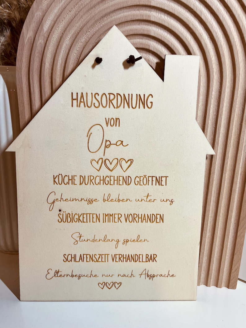 May include: A wooden house-shaped sign with the text "HAUSORDNUNG von Opa" and rules for the house, including "K&uuml;che durchgehend ge&ouml;ffnet", "Geheimnisse bleiben unter uns", "S&uuml;&szlig;igkeiten immer vorhanden", "Stundenlang spielen", "Schlafenszeit verhandelbar", and "Elternbesuche nur nach Absprache".