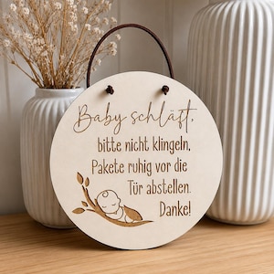 Könnte beinhalten: Ein rundes Schild aus hellem Holz mit einem dunkelbraunen Lederband zum Aufhängen. Das Schild trägt die eingravierte Aufschrift: Baby schläft, bitte nicht klingeln, Pakete ruhig vor die Tür abstellen. Danke! sowie eine Grafik eines schlafenden Babys.