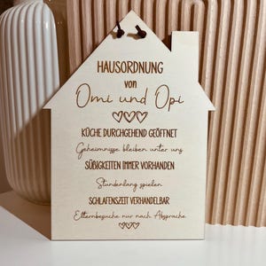 May include: A wooden house-shaped sign with the text "HAUSORDNUNG von Omi und Opi" and a list of rules: "K&Uuml;CHE DURCHGEHEND GE&Ouml;FFNET", "Geheimnisse bleiben unter uns", "S&Uuml;BIGKEITEN IMMER VORHANDEN", "Stundenlang spielen", "SCHLAFENSZEIT VERHANDELBAR", "Elternbesuche nur nach Absprache".