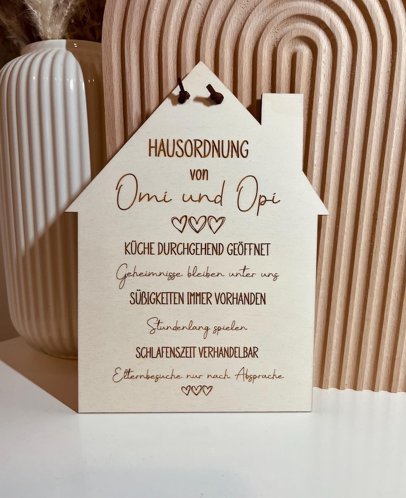 May include: A wooden house-shaped sign with the text "HAUSORDNUNG von Omi und Opi" and a list of rules: "K&Uuml;CHE DURCHGEHEND GE&Ouml;FFNET", "Geheimnisse bleiben unter uns", "S&Uuml;BIGKEITEN IMMER VORHANDEN", "Stundenlang spielen", "SCHLAFENSZEIT VERHANDELBAR", "Elternbesuche nur nach Absprache".