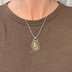 May include: A silver chain necklace with a pendant featuring a brown and yellow agate stone set in a silver bezel. The pendant has a beaded border.