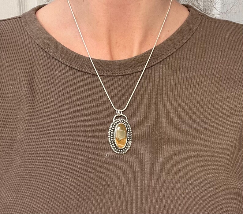 May include: A silver chain necklace with a pendant featuring a brown and yellow agate stone set in a silver bezel. The pendant has a beaded border.