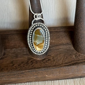 May include: A silver pendant with a brown and white agate stone set in a beaded oval frame. The pendant is hanging from a silver chain.