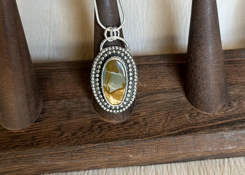 May include: A silver pendant with a brown and white agate stone set in a beaded oval frame. The pendant is hanging from a silver chain.