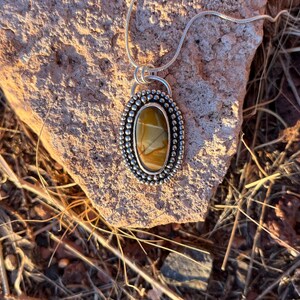May include: A silver pendant necklace with a brown and white oval stone set in a silver bezel. The stone has a unique pattern and is surrounded by small silver beads.