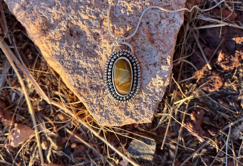 May include: A silver pendant necklace with a brown and white oval stone set in a silver bezel. The stone has a unique pattern and is surrounded by small silver beads.
