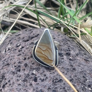 May include: A silver ring with a large, triangular stone. The stone features a pattern of tan and brown hues, resembling a landscape. The ring's setting has a decorative, textured border, showcasing a unique, handcrafted design.