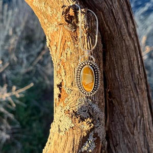 May include: A silver pendant necklace with a yellow and brown oval stone set in a silver bezel. The pendant is hanging from a silver chain and is displayed against a wooden background.