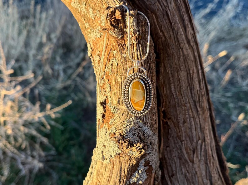 May include: A silver pendant necklace with a yellow and brown oval stone set in a silver bezel. The pendant is hanging from a silver chain and is displayed against a wooden background.
