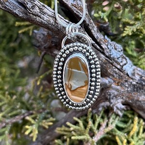 May include: A silver pendant with a brown and white agate stone set in a beaded oval frame. The pendant is hanging from a silver chain.