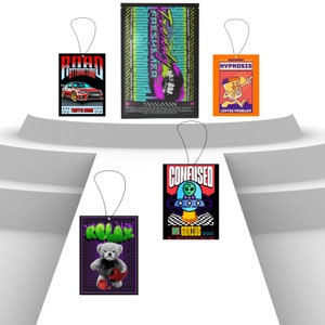 May include: Four car air fresheners with different designs. The first is black with a red car and the text "Road Attraction". The second is black with a pink and green design and the text "Trendy Freshener". The third is orange with a cartoon character and the text "Highway Hypnosis Coffee Problem". The fourth is black with a UFO and the text "Confused Off Genius".