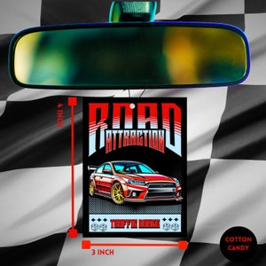 May include: A black car air freshener with a red and white design. The air freshener features a red sports car with the text "ROAD ATTRACTION" and "TRAFFIC DANCE" in white. The air freshener measures 3 inches by 4 inches.