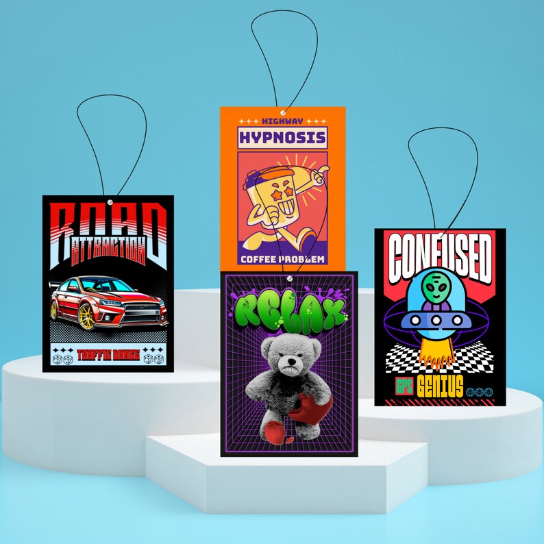 May include: Three car air fresheners with different designs. The first is black with a red car and the text "Road Attraction" and "Traffic Noise". The second is orange with a cartoon coffee cup and the text "Highway Hypnosis" and "Coffee Problem". The third is black with a green alien in a UFO and the text "Confused" and "Ops Genius".