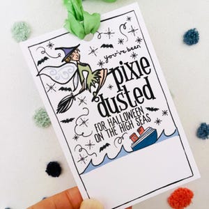 May include: A white card with a black and green illustration of a fairy flying on a broomstick. The text on the card reads "You've been pixie dusted for Halloween on the high seas".